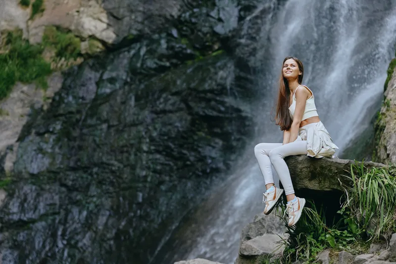 young woman sitting near waterfall in casual white clothes exploring nature outdoors on a sunny day with green foliage and rocky background