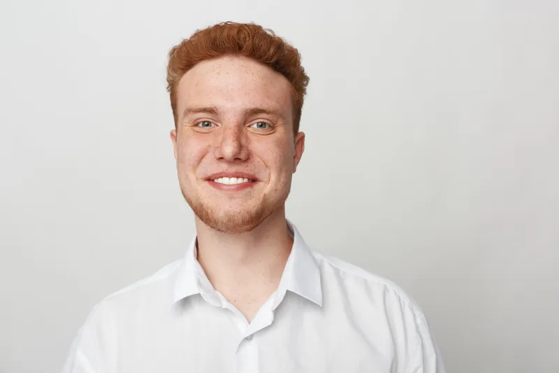 Smiling young man with red hair and beard wearing white shirt looks at camera on light background. Portrait of confident male model with natural expression and casual style in studio lighting.
