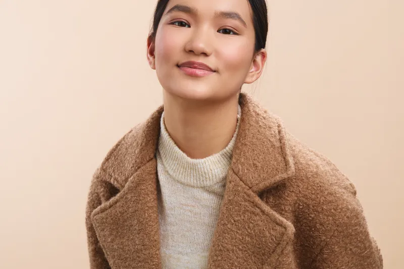 Asian woman in a cozy, stylish coat smiles softly toward the camera, conveying warmth and approachability in a simple portrait with a neutral backdrop.