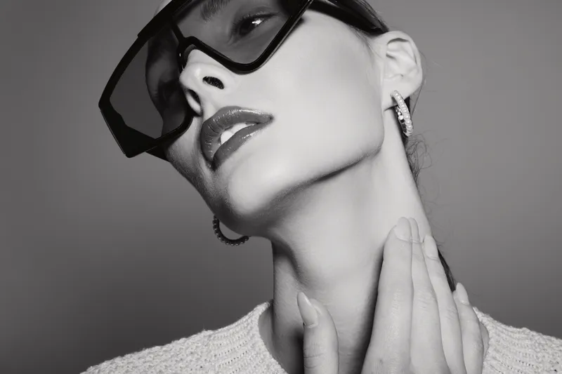 Stylish young Caucasian woman with pale skin wearing large sunglasses and hoop earrings in black and white portrait. She touches her neck with manicured hand, fashion concept.