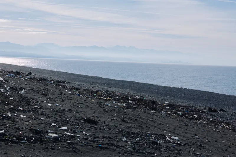 Black sand beach with litter along the shoreline, debris scattered on the coast under calm sea and distant mountains, creating a stark marine landscape.