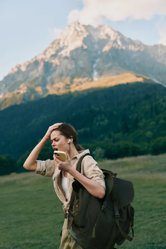 Young woman with a backpack checks her phone in a grassy meadow with towering mountains behind her, showing focus and a hint of concern.