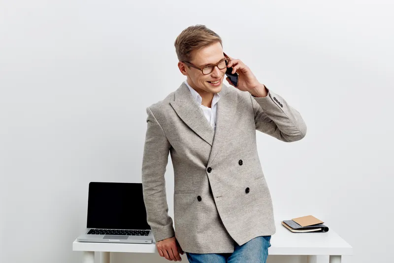 Businessman talking on mobile phone while standing in office with laptop and notepad, dressed in smart casual blazer, smiling and confident, modern work environment, professional business concept.