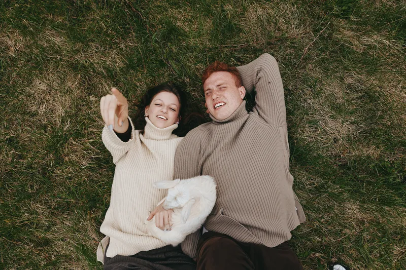 Cheerful outdoor couple lying on grass, sharing a warm blanket, wearing cozy sweaters, sunlight and breeze soften the moment, relaxed mood, natural colors, intimate connection in a simple lifestyle