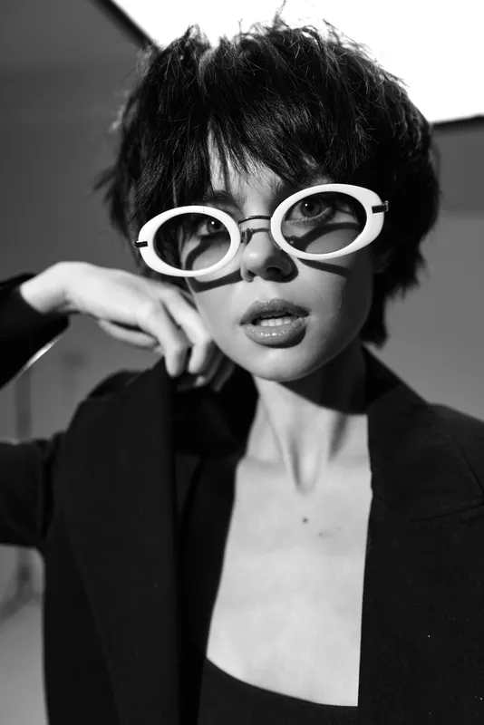 Black and white portrait of a stylish woman wearing white sunglasses with short hair and a dark jacket.