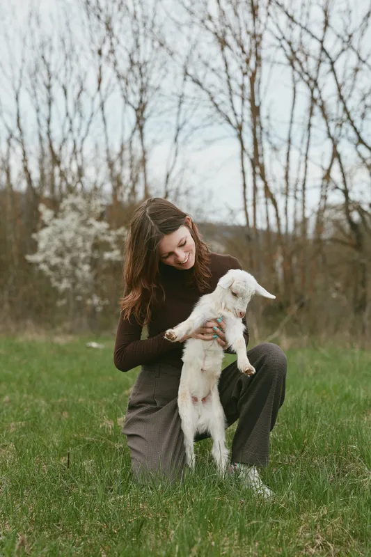 Young woman kneels on grass in a natural setting while gently holding a small white baby goat with a joyful expression on her face during a spring day.