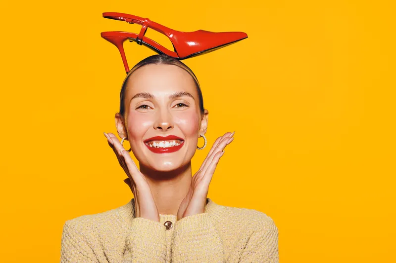 Smiling woman with red lipstick holds red high heel shoes on her head against bright orange background. Concept of fashion, beauty, fun, and creative portrait with vibrant colors and joyful mood.