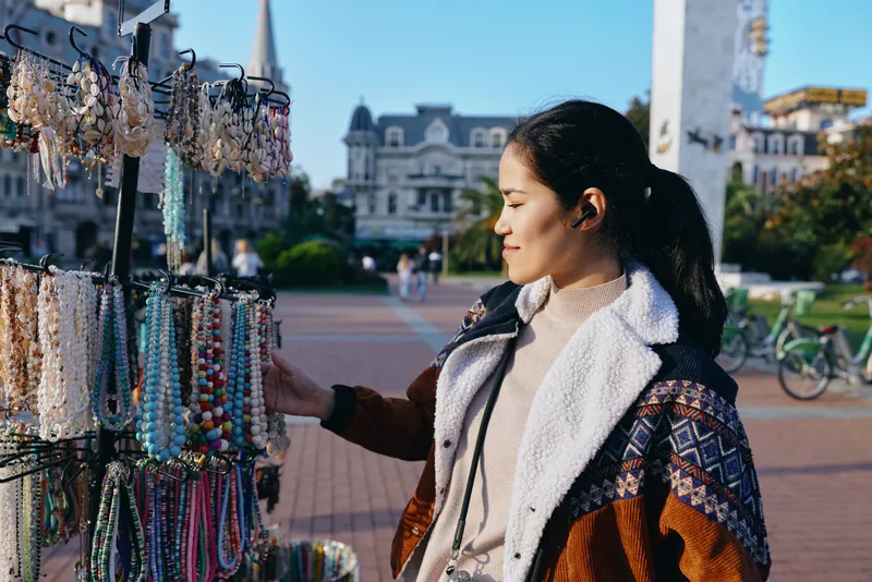 Jewelry, necklaces and colorful accessories on display at an outdoor market as a stylish woman browses bright stalls in a sunny urban square, showcasing street fashion and shopping moments