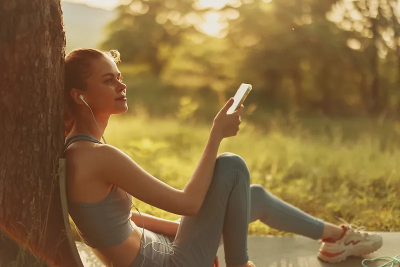 Young woman enjoying music outdoors, listening to phone with earbuds, relaxed in nature, wearing sporty attire