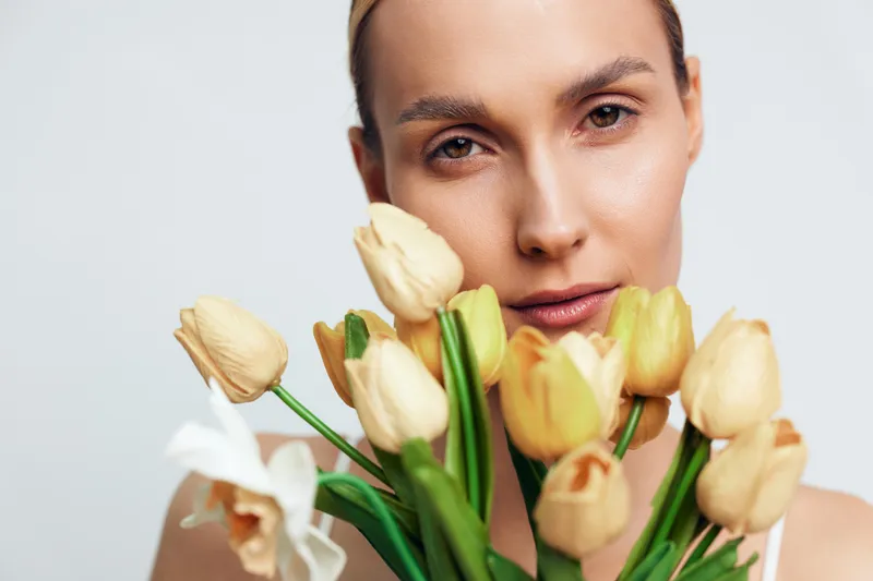 A young woman with natural makeup holds a bouquet of yellow tulips close to her face against a plain background. The image conveys freshness and spring beauty concept.