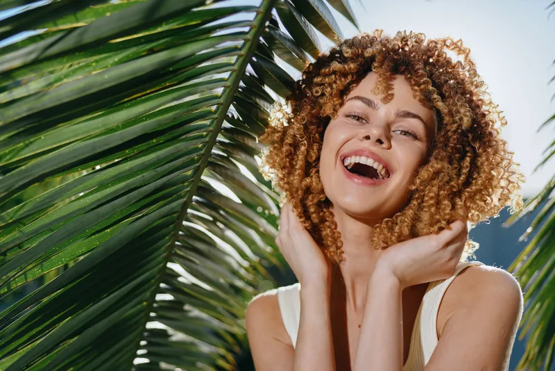 smiling woman with curly hair enjoying sunlight near tropical palm leaves, bright natural light, relaxed and happy expression, summer vibes outdoors