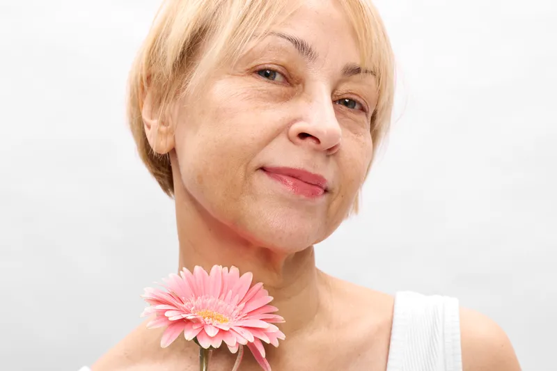Woman portrait closeup with blonde senior smile, holding a pink flower, soft white background, calm mood, gentle skincare and wellness concept