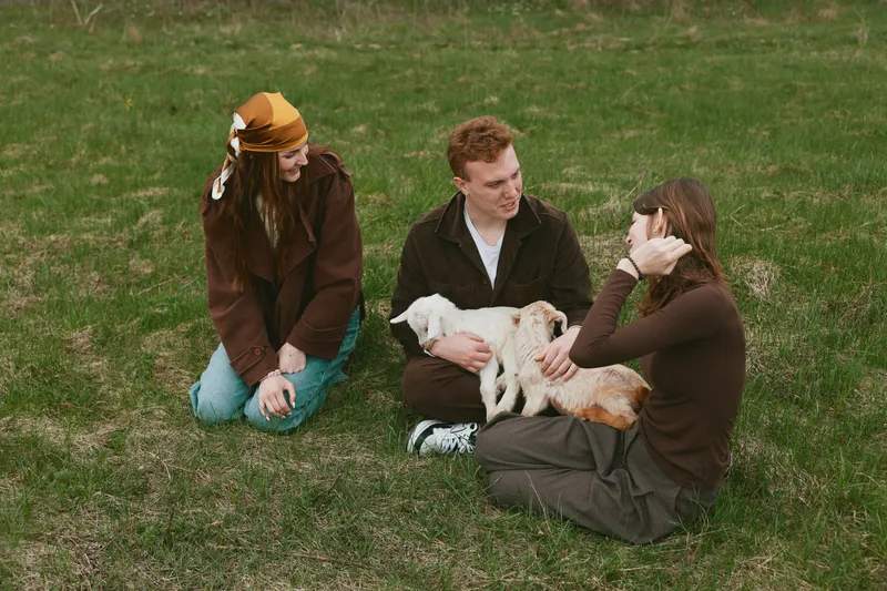 Three young adults relax outdoors on grass while holding and petting a baby goat, enjoying a casual lifestyle moment in comfortable autumn clothing.
