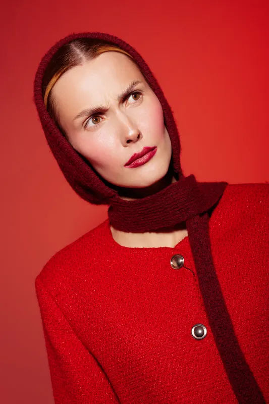 Portrait of a serious woman wearing a red coat and matching scarf on a red background. The model looks thoughtful with furrowed brows, creating a dramatic and intense mood.