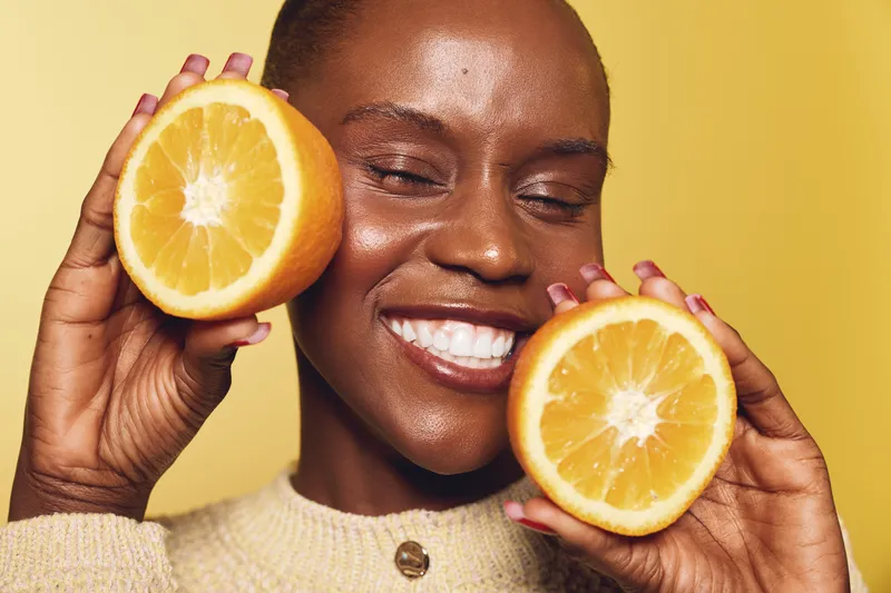 Smiling dark skinned African woman holds two halves of fresh orange close to face on yellow background. Concept of healthy lifestyle, natural beauty, vitamin C and joyful mood.
