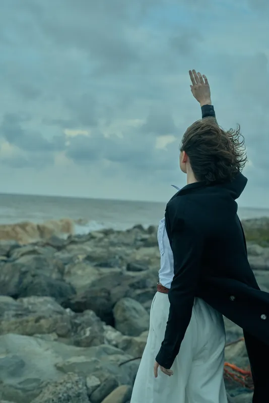Shore, ocean, wind, raised hand, dress, coat, outdoor, dramatic sky, seascape depict a lone figure at a rocky coast enjoying the windy moment and vast horizon, conveying resilience, freedom and