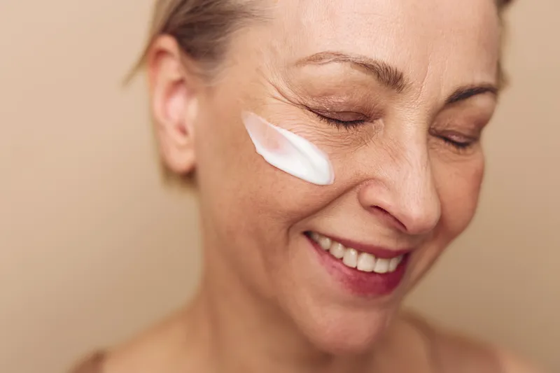 Close-up of smiling mature woman with cream on cheek applying skincare product. Concept of facial care, anti aging, beauty routine and natural skin health in soft warm lighting.