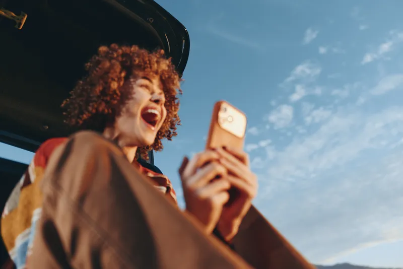 Outdoors lifestyle portrait of a woman with curly hair laughing, holding a smartphone for selfie, summer sky behind, bright daylight travel vibe, candid street moment.