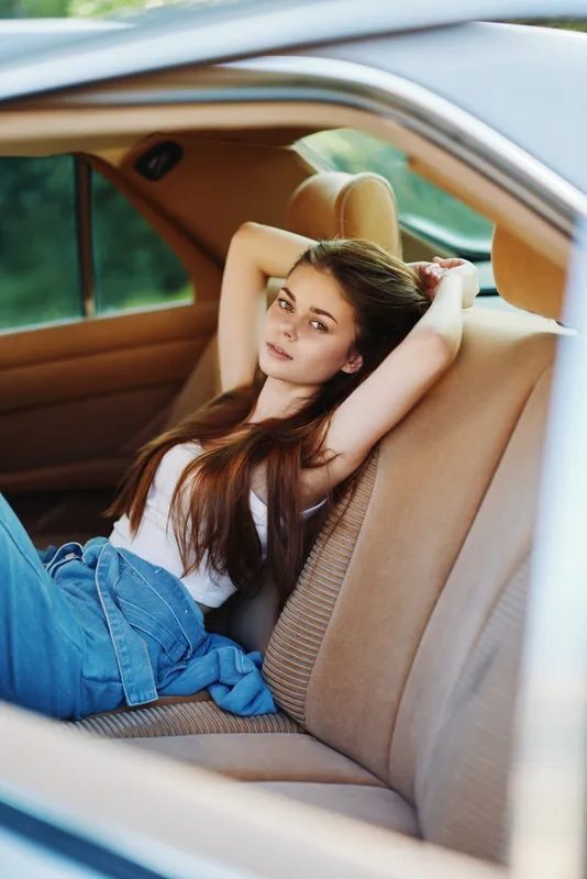 Stylish young woman with long hair relaxing in a car, showcasing casual summer fashion with denim and soft emotions in a nature-inspired setting.