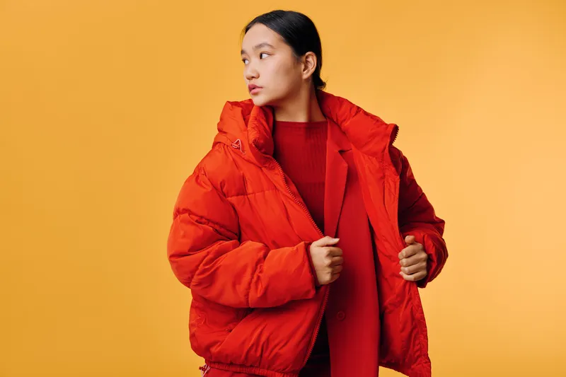 Woman wearing bright red winter coat and sweater standing against yellow background looking sideways. Fashion, warm clothing, casual style and cold weather outerwear for fall or winter season.