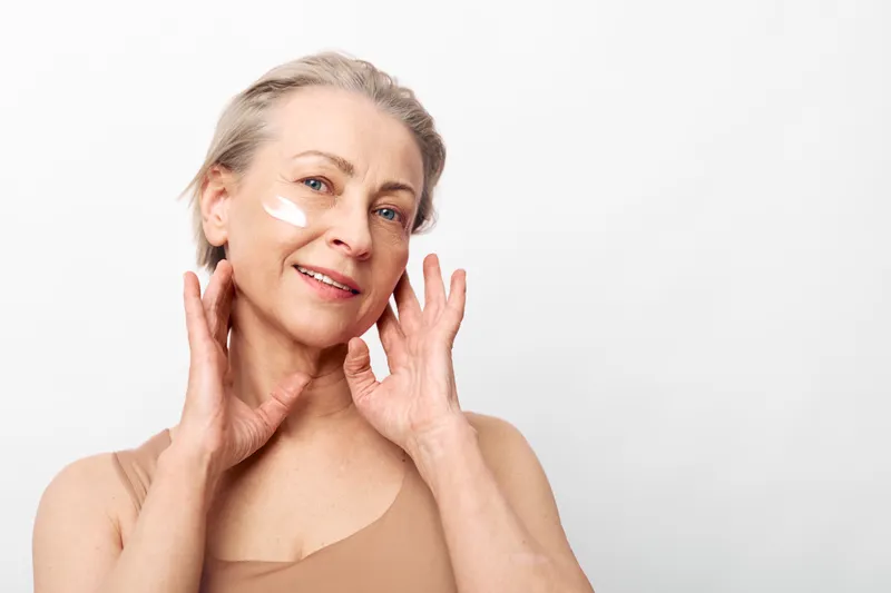 Mature woman with light skin applies moisturizing cream on face, smiling gently in bright studio. Concept of skincare, aging beauty, self-care, and natural wellness at home.