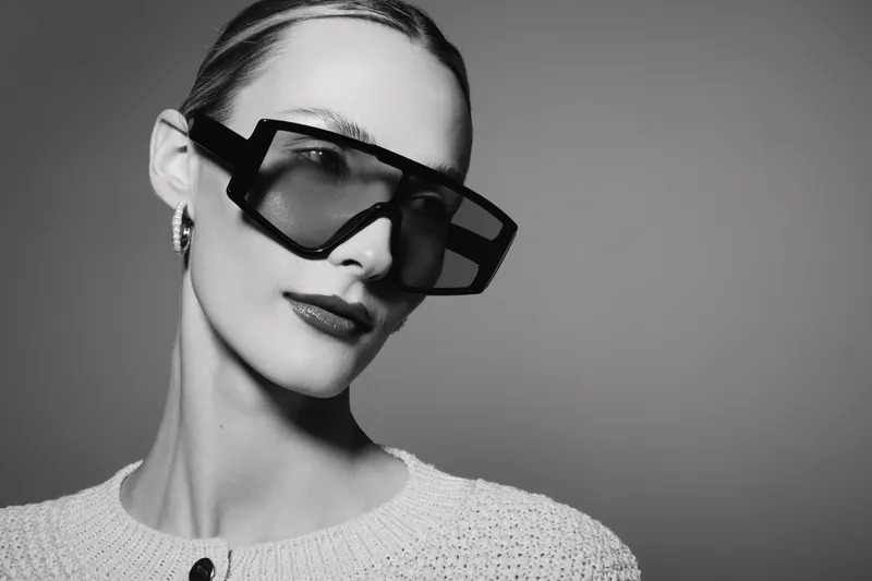 Stylish Caucasian woman with fair skin wears oversized sunglasses and white knitted top posing with confident expression in studio. Fashion and elegance concept.