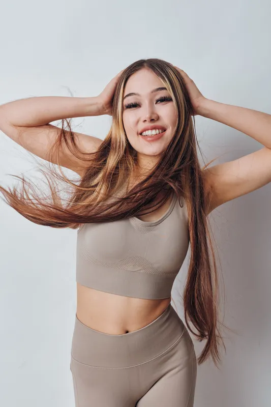 Young woman with long hair smiling and posing with hands on head in beige sportswear. Studio portrait of happy girl with natural makeup and flowy hair against white background.