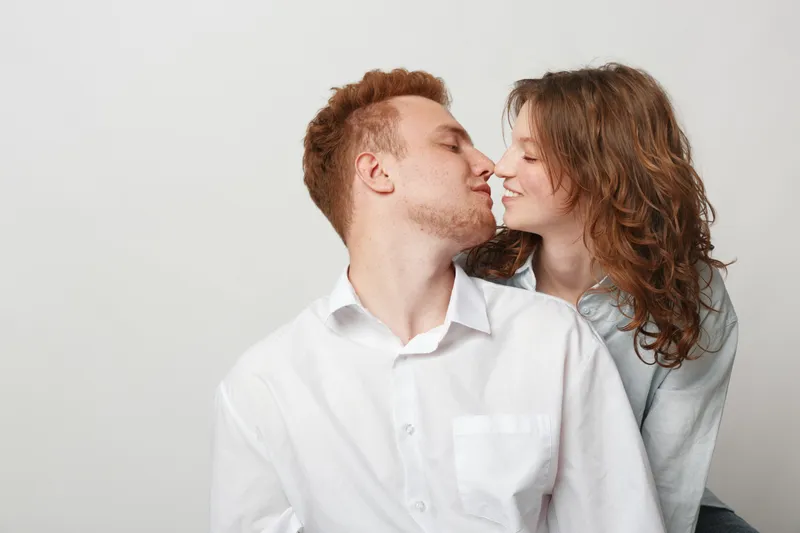 Young couple in casual clothes shares intimate moment, leaning close with eyes closed. Light background highlights their affectionate connection and tender emotions in a cozy setting.