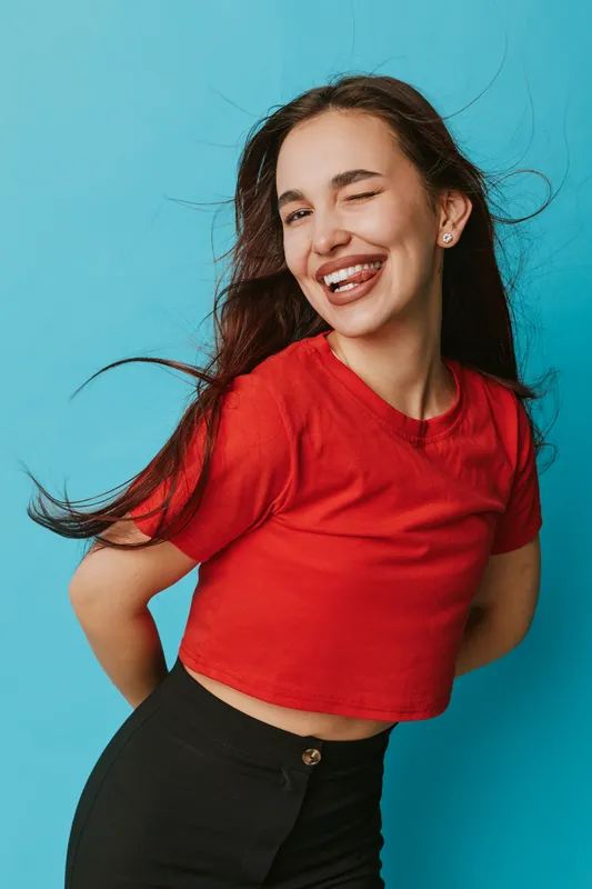 Young woman with long hair wearing red crop top and black pants winking and smiling playfully on blue background. Casual fashion, happiness and youthful energy with confident pose.