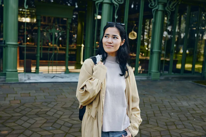 Woman wearing beige coat and gray sweater stands outdoors near a green storefront, casual urban style, thoughtful pose, daytime stroll, everyday fashion photography scene