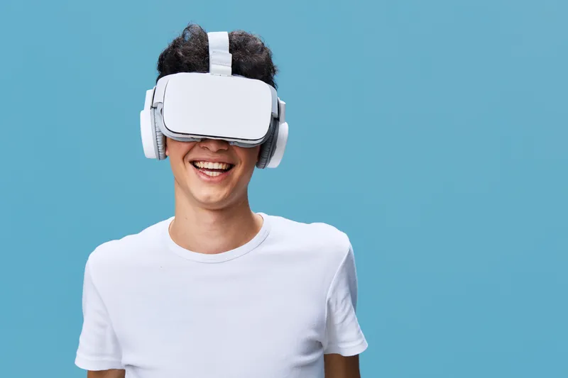 Young man wearing virtual reality headset and wireless headphones smiling joyfully against blue background, experiencing immersive technology and digital innovation.