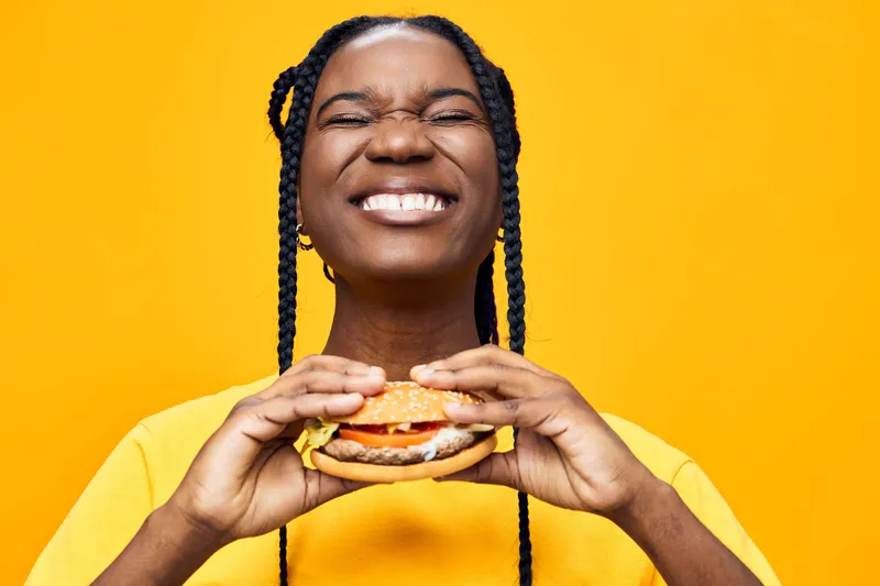 Happy woman holding a delicious burger in front of a vibrant yellow background, showcasing her joy and love for food, perfect for healthy eating concepts