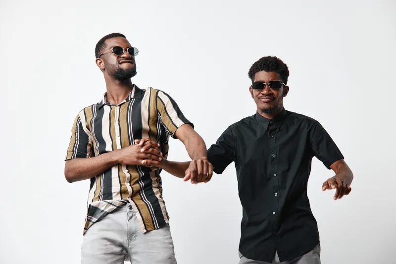Two young black men with dark skin wearing sunglasses and casual shirts dancing and having fun together on white background. Friendship, joy and style in studio.