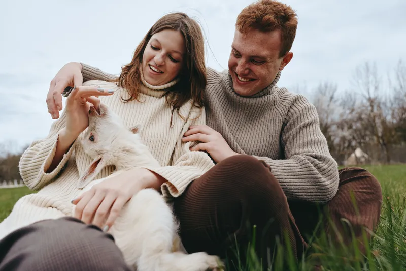 A smiling couple sits on a grassy outdoor patch, gently stroking a friendly goat kid. A cozy lifestyle moment captures affection, connection, and simple countryside joy in daylight.