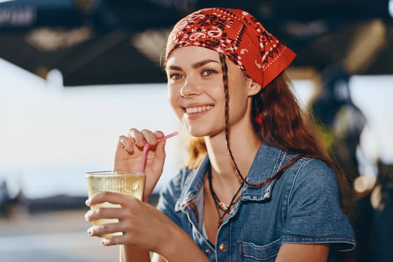 smiling woman in modern boho-western denim street style drinking beverage, wearing red bandana, film color, casual outdoor summer lifestyle portrait