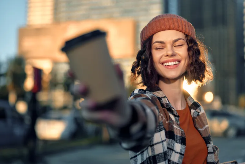 Woman holding coffee smiling in city street, candid urban moment at golden hour with beanie and backlight, authenticity and candid lifestyle captured in joyful emotional storytelling.