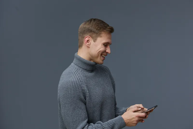 Smiling man wearing a gray turtleneck sweater using his smartphone, holding it with both hands, isolated on a plain gray background, studio portrait, casual style, happy mood, modern technology.