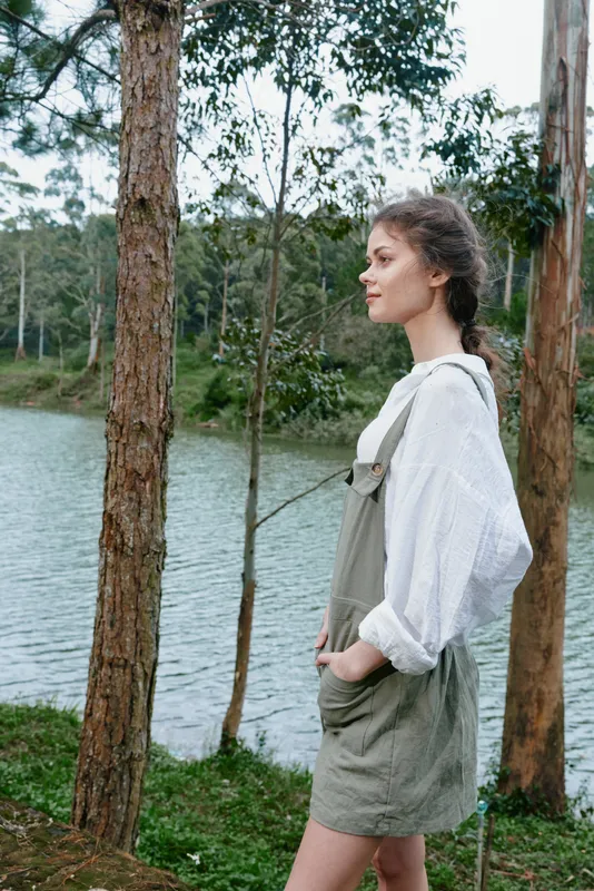 Young woman in casual outfit by the lake, expressing serene emotions, surrounded by nature and trees, evoking a peaceful summer vibe with green tones