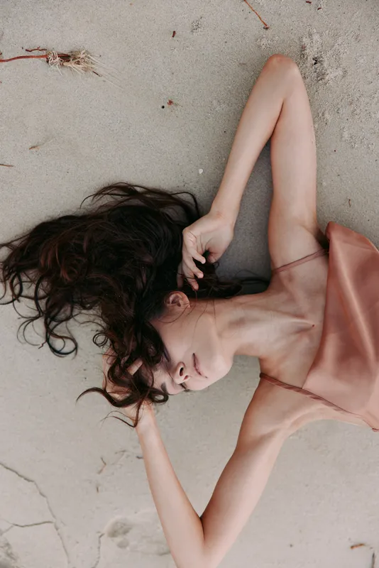 A woman rests on the sandy ground with eyes closed, dark hair spread across the shore, and a soft pink dress draped over her body, capturing serene coastal stillness.