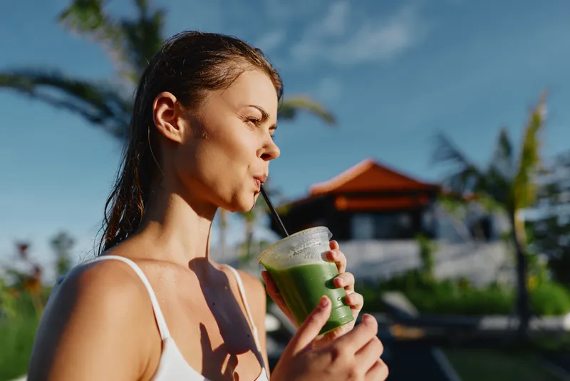 A lifestyle shot of a woman enjoying a green smoothie outdoors in bright sun, a relaxing vacation vibe and health focus, emphasizing hydration, wellness, and self care in a sunny, carefree moment.