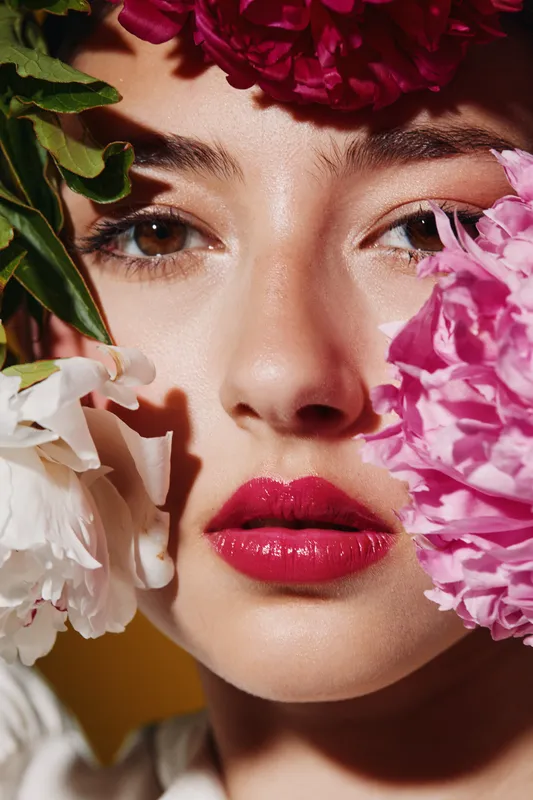 Close-up of a young woman with glossy red lips surrounded by vibrant flowers. Her smooth skin contrasts with the bright petals. Concept of beauty, creative, and fashion portrait.