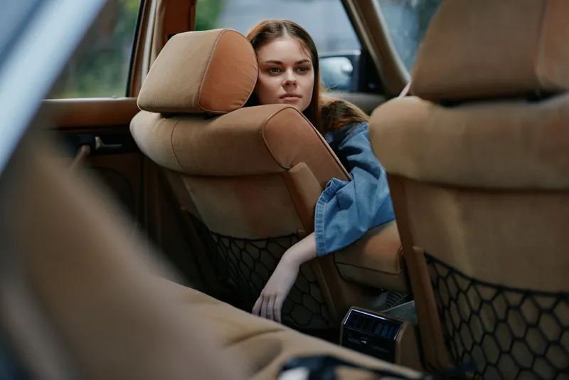 Young woman sitting in a vintage car, contemplating life with a thoughtful expression, dressed casually in a denim outfit, surrounded by soft brown upholstery.