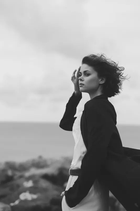 Woman walking along a windswept coastline in monochrome, hair blown by wind, contemplative mood, casual outfit, ocean horizon behind.