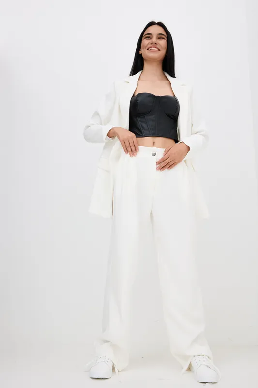 tall woman in white suit and black corset stands confidently with relaxed pose, smiling gently against plain white background in stylish modern outfit.