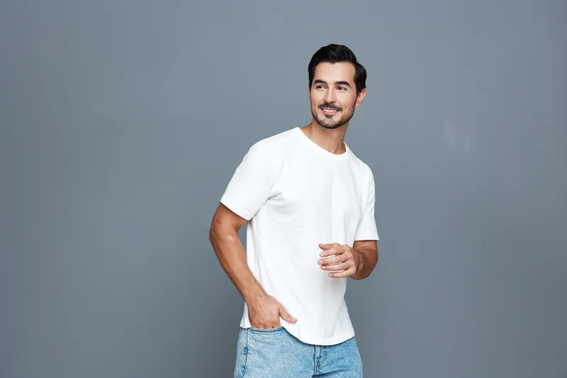 Smiling man dressed in casual white t-shirt and blue jeans stands confidently against a plain gray background, showcasing a relaxed and approachable vibe.