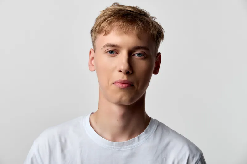 Casual young man with short blond hair in a white t shirt facing the camera with a neutral expression, soft studio lighting and a simple background for fashion or profile use