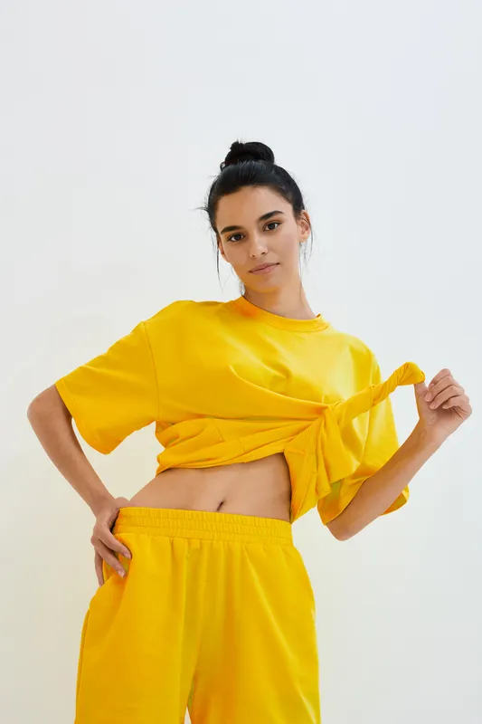 Young woman wearing bright yellow casual outfit poses confidently against a plain white background, showcasing a sporty and relaxed style with vibrant colors.
