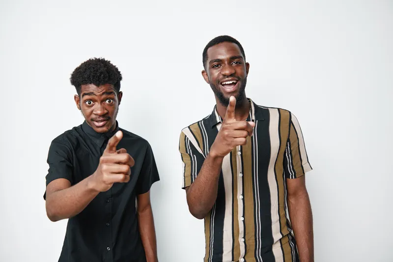 Two african men with dark skin pointing at camera smiling and standing together in casual clothes on white background. Young black males with happy expressions and friendly gesture.