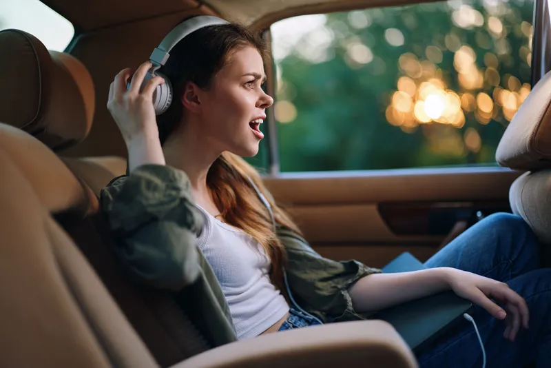 Young woman enjoying music in a car, dressed in casual wear, expressing joy, surrounded by natural light and greenery during sunset.