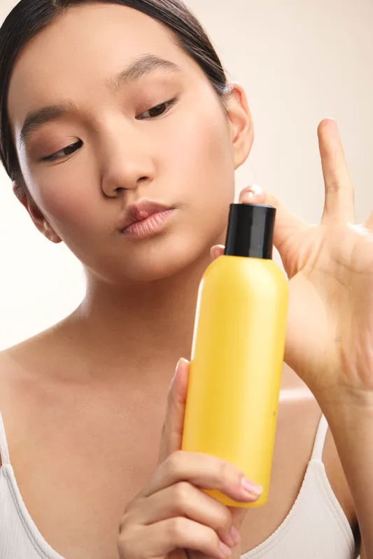 asian woman examines a yellow bottle with a pump in a bright, minimal setting, preparing a skincare routine and exuding calm focus and selfcare.